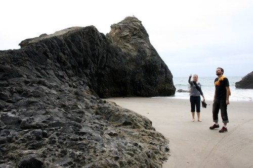 Two people stand on a sandy beach near large rocky formations, with the ocean in the background under a cloudy sky.