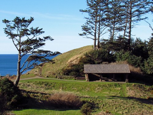 A coastal landscape featuring a grassy area, a wooden shelter, and trees overlooking the ocean under a clear blue sky.