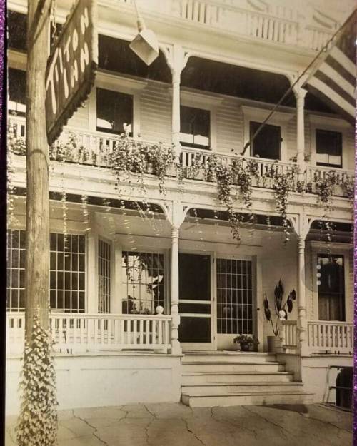 Historic photo of a two-story inn with a sign, porch, and decorative plants, showcasing classic architecture.