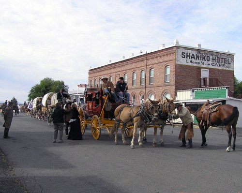 A historic street scene with a horse-drawn carriage, wagons, and people in period costumes near a brick hotel.