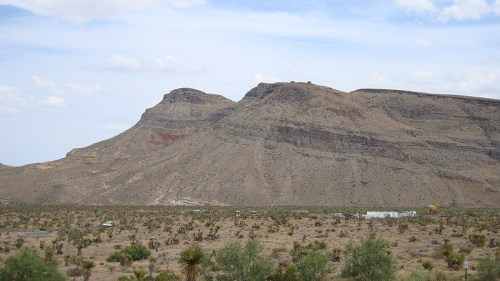 A rugged mountain landscape with a flat-topped peak, surrounded by sparse desert vegetation under a cloudy sky.