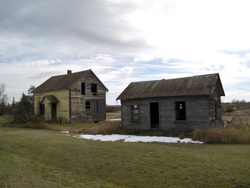 Two abandoned houses, one yellow and one gray, sit side by side in a grassy field under a cloudy sky.