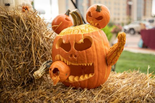A creatively carved pumpkin with a fierce face, holding a smaller pumpkin, surrounded by hay bales.