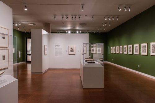 Art gallery interior featuring framed artworks on green walls, with display cases and informational panels.