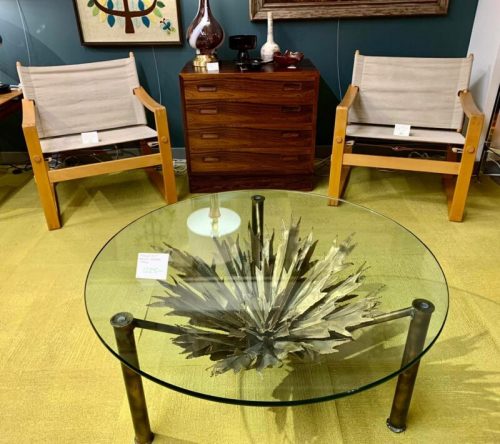 A modern living room setup featuring two wooden chairs, a glass coffee table with a starburst design, and a wooden cabinet.