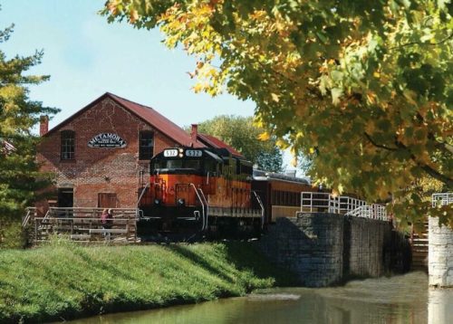 A colorful train parked by a historic brick building near a calm waterway, surrounded by trees in autumn foliage.