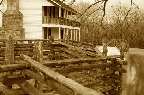 A sepia-toned image of a historic house with a wooden fence in the foreground and bare trees in the background.