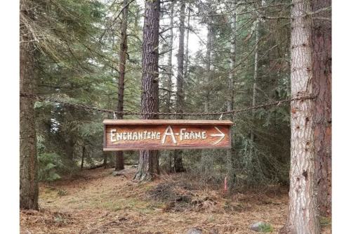 Wooden sign reading "Enchanting A-Frame" with an arrow, surrounded by tall trees in a forest.