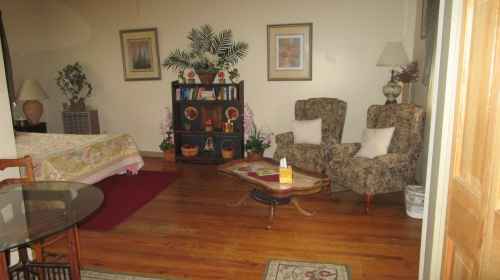 Cozy living space with two patterned chairs, a wooden table, and a bookshelf, featuring warm lighting and wooden floors.