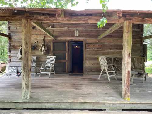 A rustic wooden cabin with a porch, featuring rocking chairs and a wheel, surrounded by greenery.