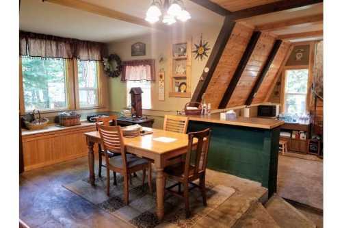 Cozy kitchen with wooden dining table, chairs, and large windows, featuring rustic decor and an A-frame ceiling.