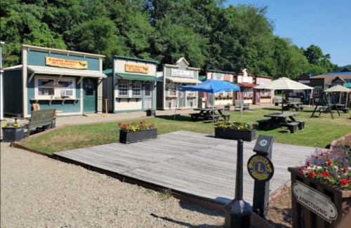 A quaint outdoor area with colorful storefronts, picnic tables, and umbrellas surrounded by greenery.