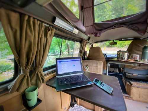 A cozy camper interior with a laptop on a table, a green mug, and a remote, surrounded by nature.