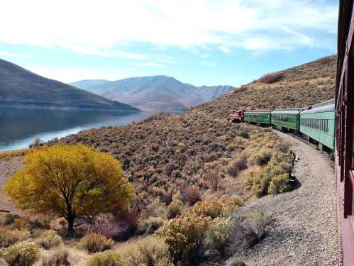 A scenic view of a train traveling along a river, surrounded by mountains and autumn foliage.