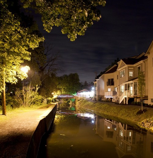 A serene night scene of a canal with reflections, lined by trees and illuminated houses under a cloudy sky.