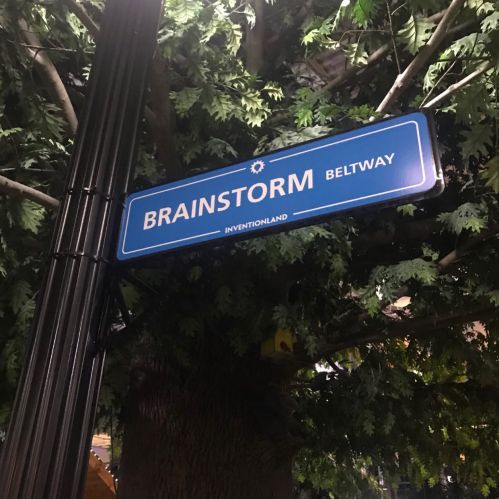 A street sign reading "BRAINSTORM BELTWAY" surrounded by green foliage.