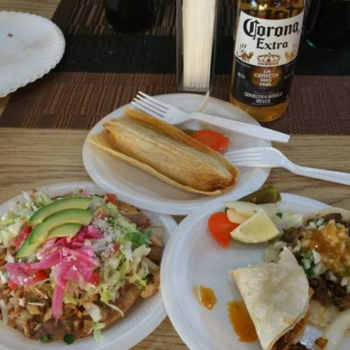A plate of tacos and tamales with toppings, accompanied by lime wedges and a bottle of Corona beer.