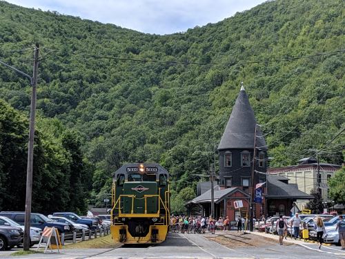 A green and yellow train stops near a historic building, with a crowd and lush green mountains in the background.