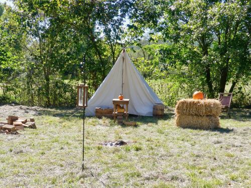 A white tent in a grassy area, with hay bales, a wooden table, and pumpkins surrounded by trees.