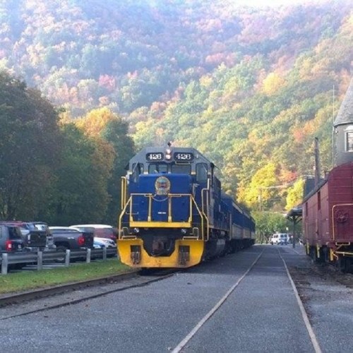A blue and yellow train on tracks with colorful autumn trees in the background and parked cars nearby.
