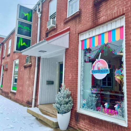 A brick building with a colorful shop window displaying a unicorn theme, surrounded by snow. Signs indicate it's open.