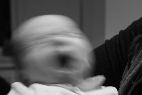 A blurred image of a baby crying, with a hand partially visible in the frame. The scene is in black and white.