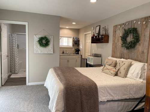 Cozy bedroom with a bed, kitchenette, and bathroom entrance, decorated with wreaths and neutral tones.
