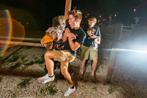 A young man interacts with a monkey while another person laughs in the background, all in a nighttime setting.