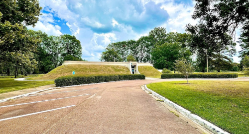 A grassy mound with a small entrance, surrounded by trees and a parking area under a partly cloudy sky.