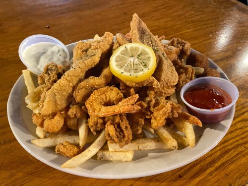 A plate of fried seafood, including shrimp and fish, served with fries and dipping sauces, garnished with a lemon slice.