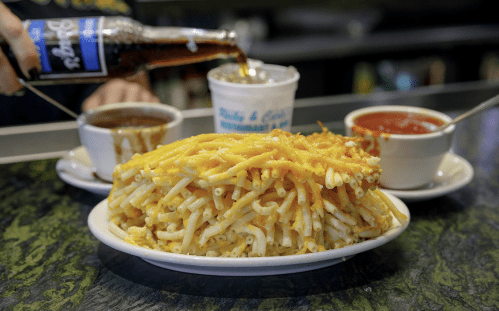 A plate of cheesy macaroni, with cups of soup and sauce, and a bottle of soda being poured into a cup.