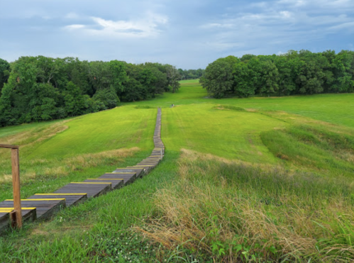 A wooden pathway leads through lush green fields, surrounded by trees under a cloudy sky.