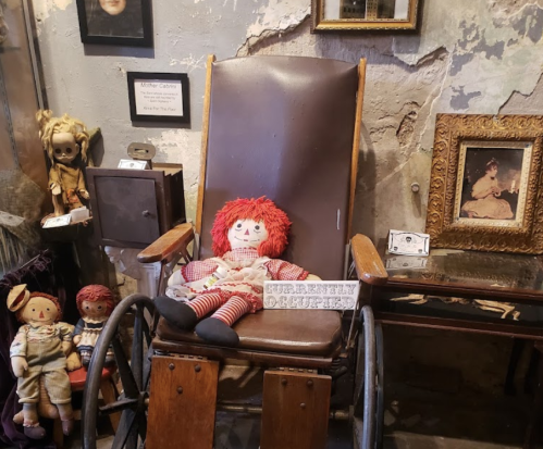 A vintage rocking chair with a red-haired doll sitting on it, surrounded by other dolls and framed photos.