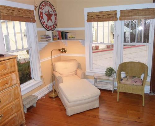Cozy corner with a chaise lounge, a lamp, and a wicker chair, surrounded by windows and warm yellow walls.
