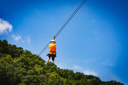 A person in an orange safety vest zip-lining over a lush green landscape under a clear blue sky.