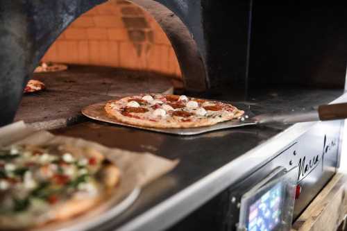 A pizza being removed from a wood-fired oven, with another pizza visible inside the oven.