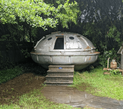 A metallic, UFO-shaped structure sits on a grassy area, surrounded by greenery and steps leading to its entrance.