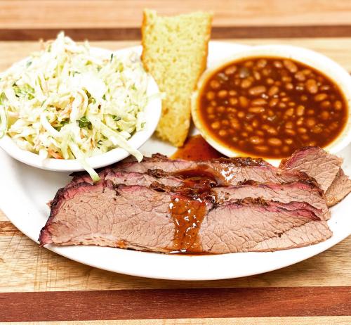 A plate featuring sliced brisket with sauce, coleslaw, baked beans, and a slice of cornbread on a wooden table.