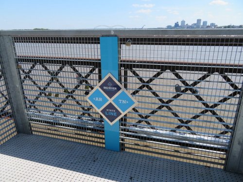 Sign marking the state line between Arkansas and Tennessee on a bridge over the river, with a city skyline in the background.