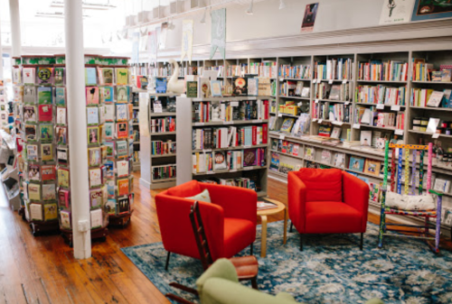 A cozy bookstore interior featuring red chairs, a wooden table, and shelves filled with books and greeting cards.