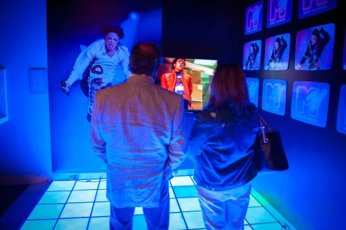 Two people watch a video of a performer in a colorful, illuminated exhibit space with blue lighting.