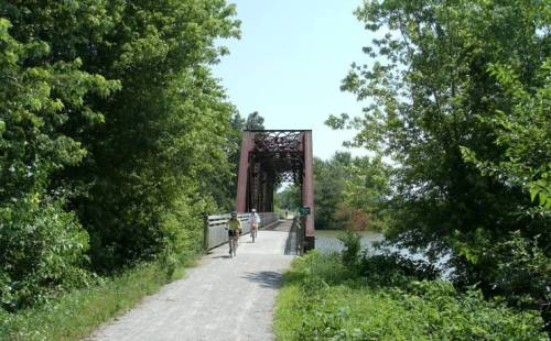 A path leads to a red metal bridge surrounded by lush green trees and a calm waterway. Two people walk along the path.