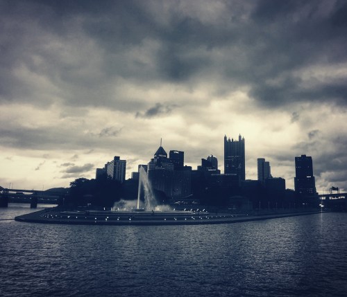 Silhouette of Pittsburgh skyline against a moody sky, featuring a fountain in the foreground and dark clouds above.