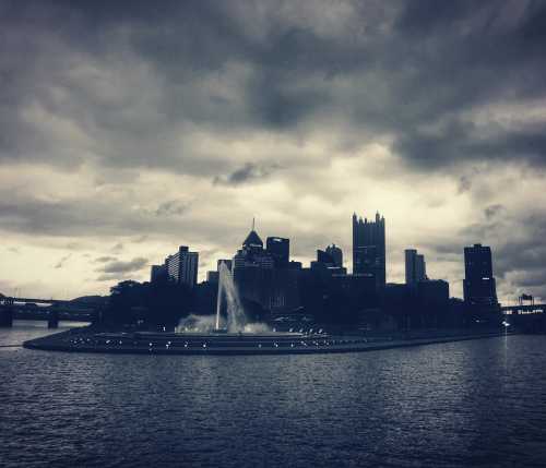 Silhouette of Pittsburgh skyline against a moody sky, featuring a fountain in the foreground and dark clouds above.