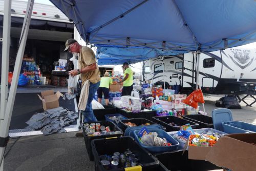 Volunteers organize food and supplies under blue tents at a community aid event, with boxes and bins around them.
