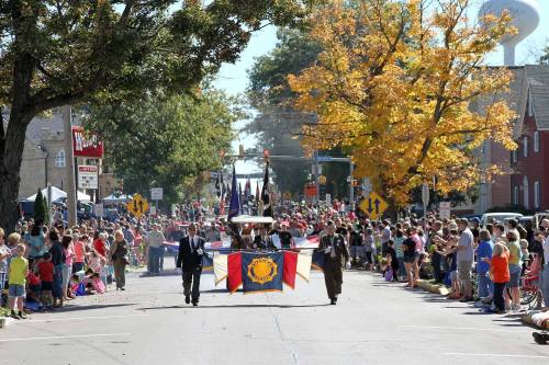 A parade scene with people lining the street, colorful flags, and autumn trees in the background.