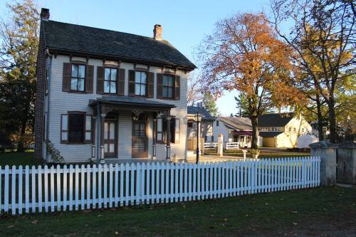 A two-story historic house with a white picket fence, surrounded by trees and other buildings in a rural setting.