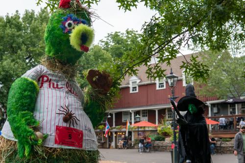 A large green mascot in a Phillies jersey stands next to a witch figure, set in a festive outdoor area.