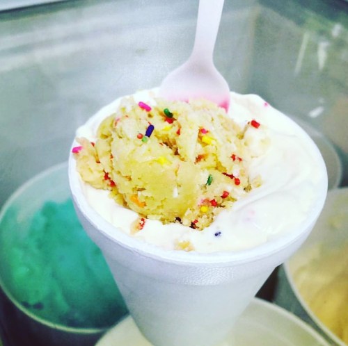 A cup of ice cream topped with colorful sprinkles and a pink spoon, set against a background of ice cream tubs.
