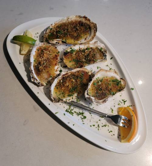 A platter of baked oysters topped with breadcrumbs, garnished with parsley, lime, and an orange slice.
